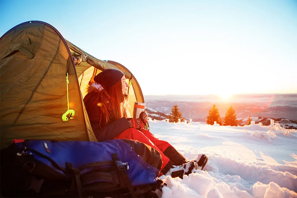A person enjoying the sunrise sitting at the entrance of a tent while holding a thermal water bottle with a bag beside on snow covered field. In the background is clear white skies with hues of yellow and a couple of trees.