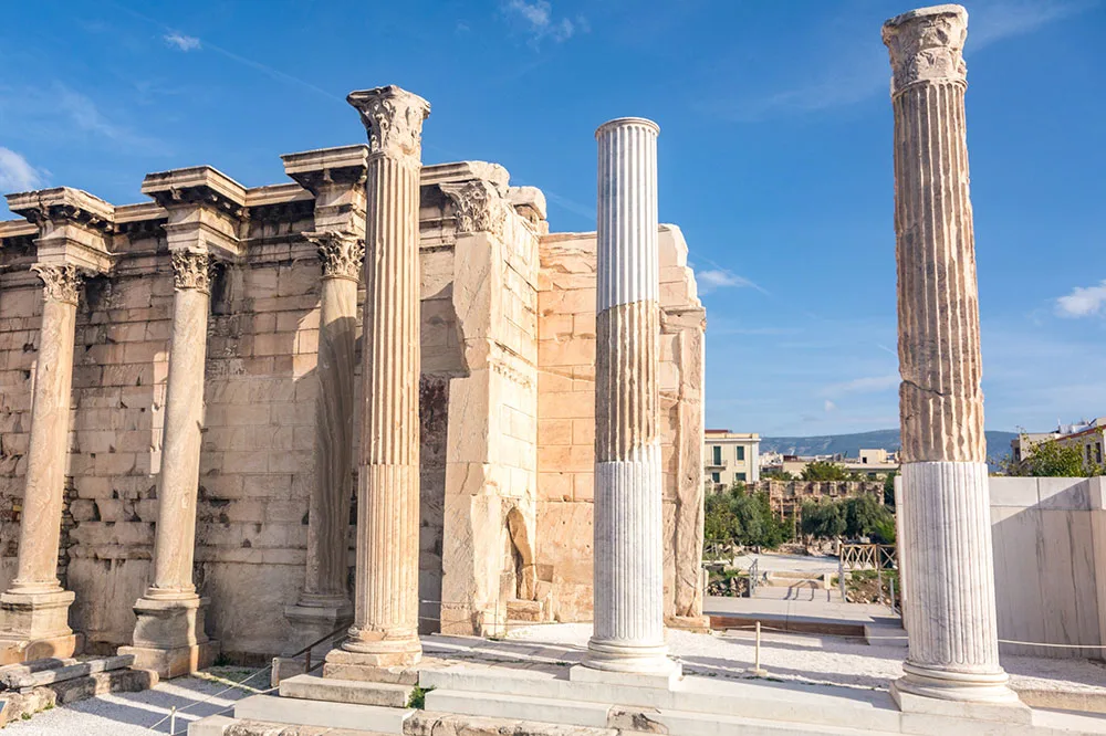 Pillars standing that are once part of a wall with a couple of pillars of the Hadrian’s Library under bright blue skies. In the back are buildings covered by trees.