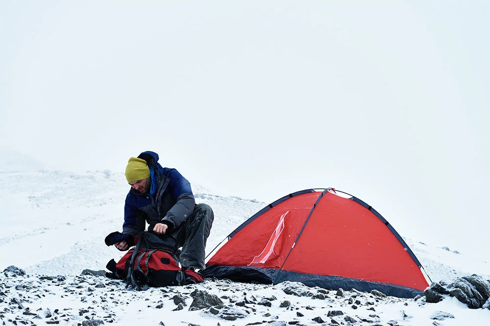 A person putting things back to the hiking bag beside a tent on a snow and stone covered field under white skies.