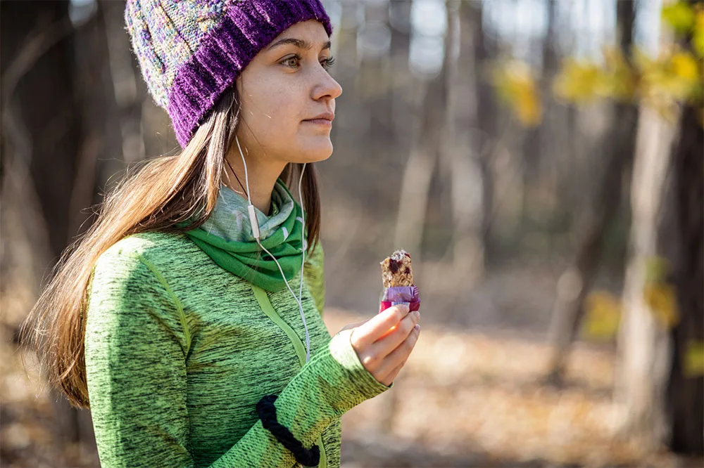 A person wearing a winter scarf, thick hat, and long-sleeved base clothing while eating high energy snacks with blurry view of trees in the background.