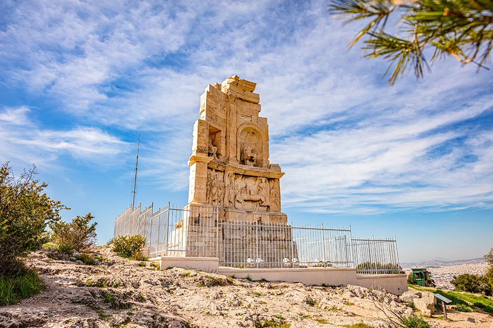 A structure in the middle filled with carvings protected by walls surrounded by dirt road and bushes under bright blue skies with scattered clouds.