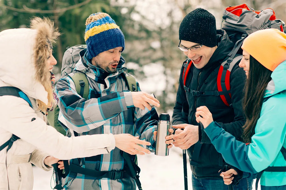 A person opening an insulated bottle with three people in hiking gear on snow covered path with a blurry view of trees in the background.