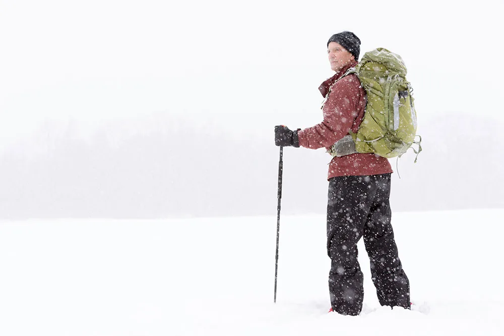 A person surrounded by snow wearing a hiking cap, green bag, and black pants while holding a trekking pole.