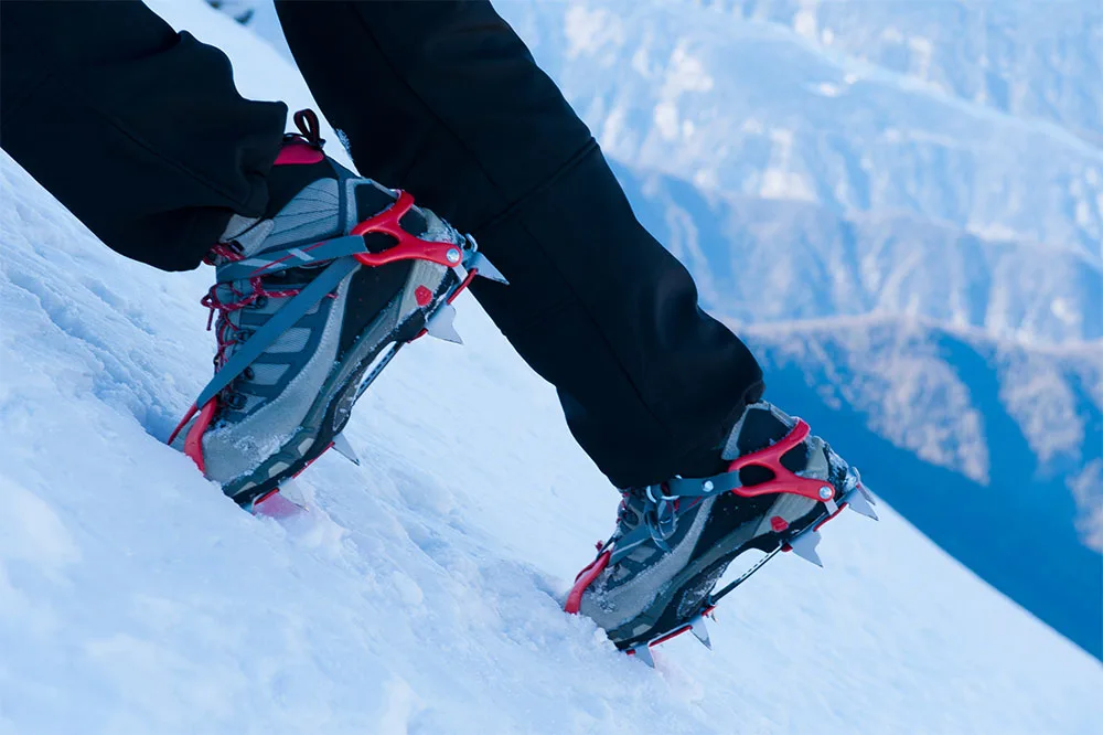A person is wearing hiking pants, and red spikey crampons on a gray and black shoes while climbing on an icy slope with mountains in the background.