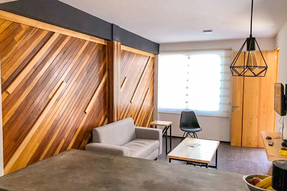 Inside a room with rustic finishings, a mixture of wooden paneled and black painted walls. A gray small sofa, two wooden tables, and a black chair can be seen in the middle.