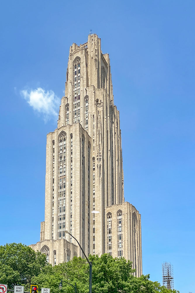 The tall white building of the Cathedral of Learning University of Pittsburgh Main Campus under clear blue skies emerging from the green trees below.