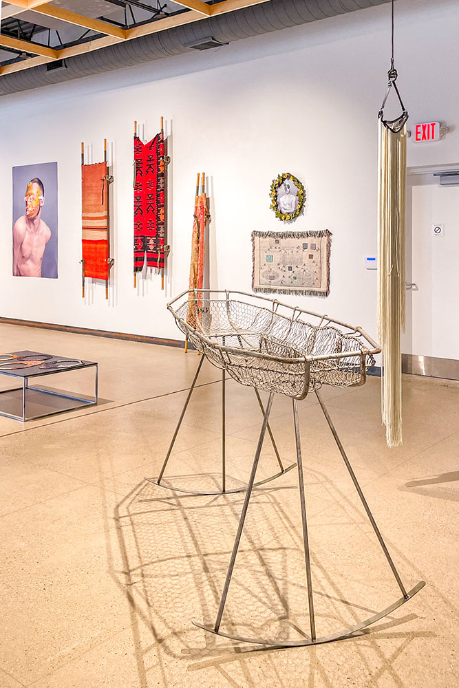 A raised vintage metal basket on a brown tiles in the center of a display pattern textile, paintings, and a table filled with colorful necklaces in a white walled room in Lawrenceville, Pittsburgh.