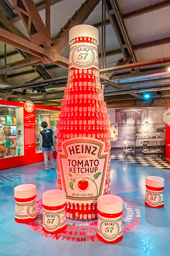 A display of Heinz bottles forming a large Heinz Tomato Ketchup bottle surrounded by the top cap of the bottles in the middle of a room with blue floors and display of the history of the ketchup brand.