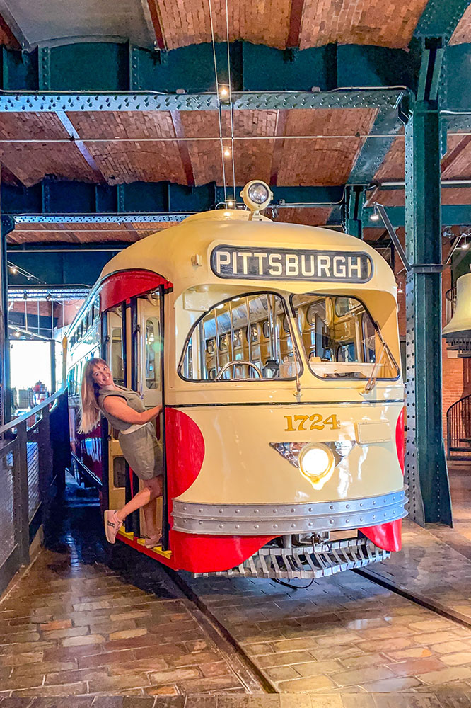 Me on the entrance of the Pittsburgh display of a white and red train inside a bricked wall room.