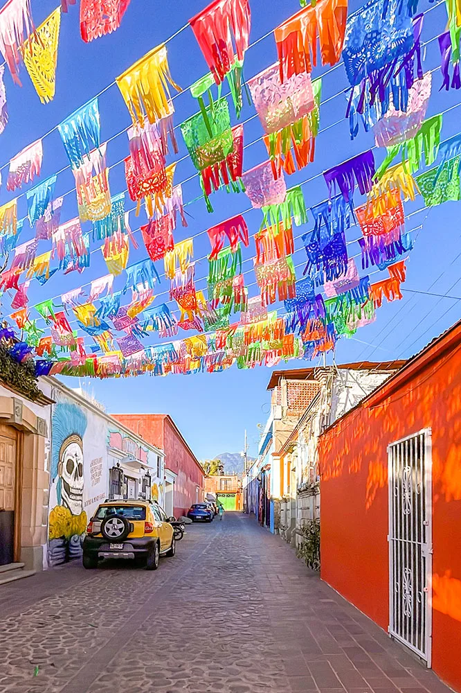 A street with cobbled stone and slate tiles with parked cars and lines of houses painted with murals and bright colors. Paper confetti are hanged across the street under clear blue skies in Oaxaca Mexico