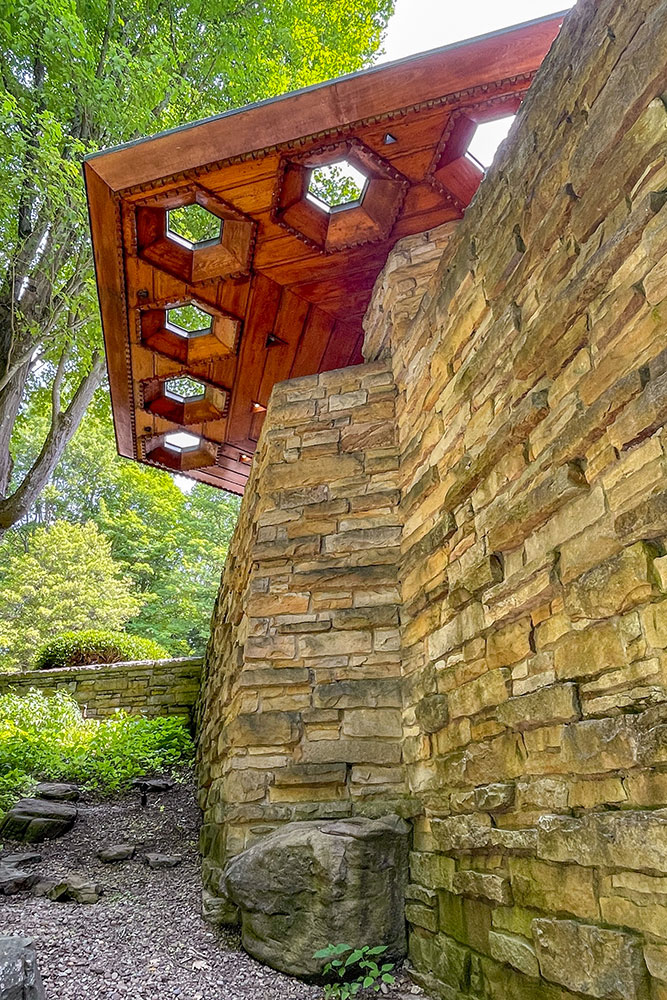 The stone wall of the Fallingwater showing the octagon sun windows of the wooden ceiling surrounded by tall trees and lush vegetation.