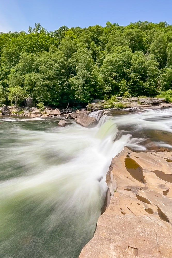 The water flowing from the Ohiopyle Falls with the lush green trees at the back under clear blue skies.