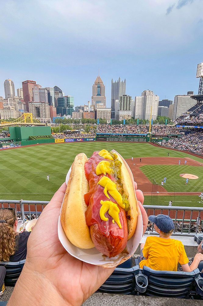 A hand holding a hotdog from a bench with people watching a baseball match in the background with a view of tall buildings under clear blue skies. 