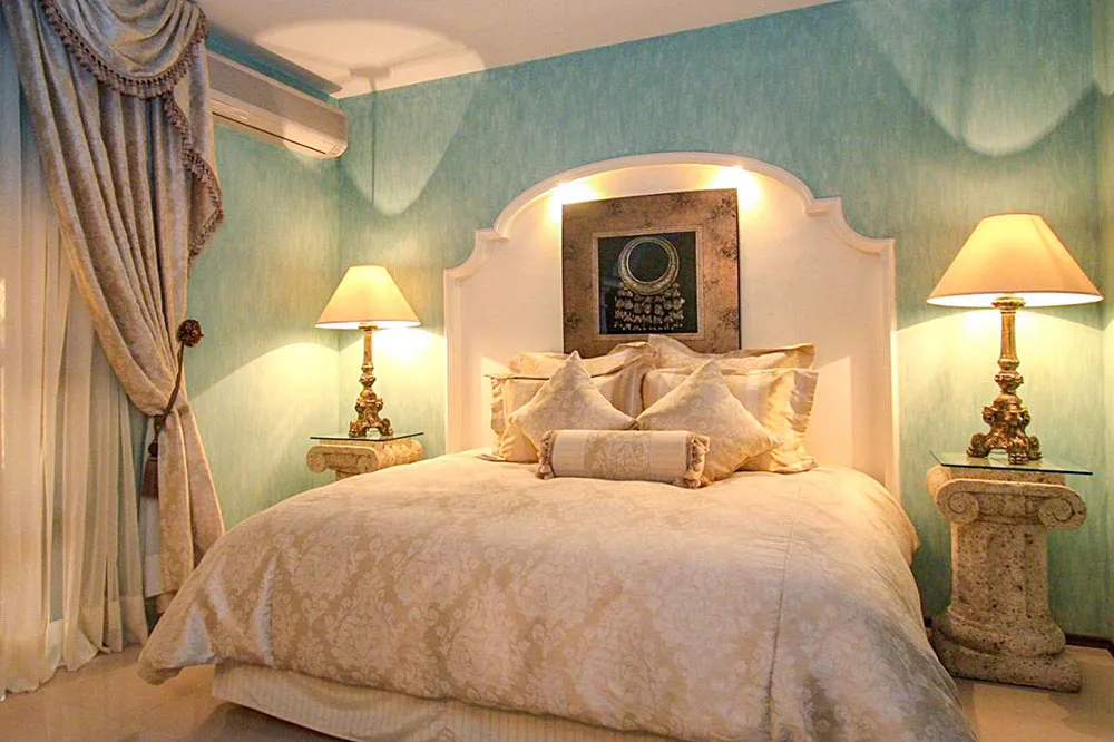 A room with sky blue walls with a queen sized white bed with gold accents and a white tall headboard with two side tables with light lamps, a best place to stay in Oaxaca