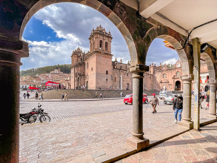 A wall with arched open walls showing an outdoor cobbled stone space featuring a parked red car and people walking. In the background shows the side view of the Cusco Cathedral under bright cloudy skies to visit in 2 days in Cusco.