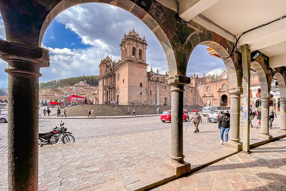 A wall with arched open walls showing an outdoor cobbled stone space featuring a parked red car and people walking. In the background shows the side view of the Cusco Cathedral under bright cloudy skies to visit in 2 days in Cusco.