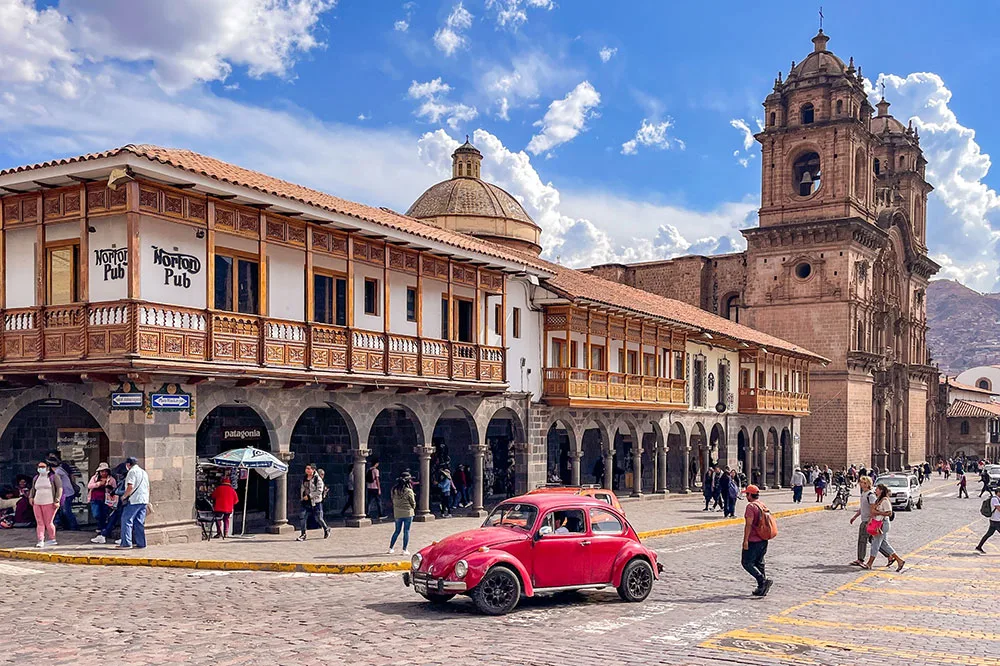A cobblestone road with a red car in the middle and few people walking with a long 2 level white building in the Cuzco Main Square