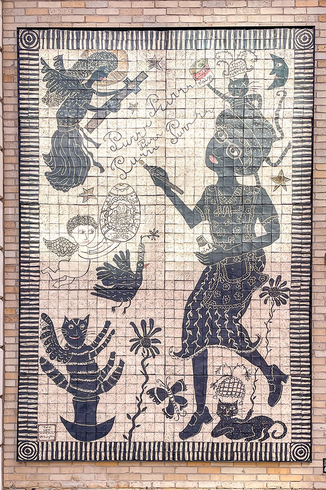 A black and white tiled artwork of angels, cats, and a woman writing on bricked wall of a building in Pittsburgh. 