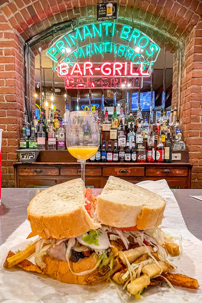 Overflowing sandwich, fries, and veggies on a gray table, paired with a drink in a wine glass. Display of bottles, glass wall, and neon sign.