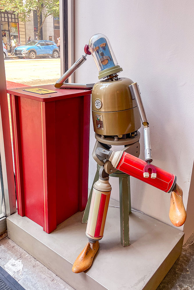 A robot installation leanign on a tall red table on a corner with white wall and floor to see in a weekend in Pittsburgh.