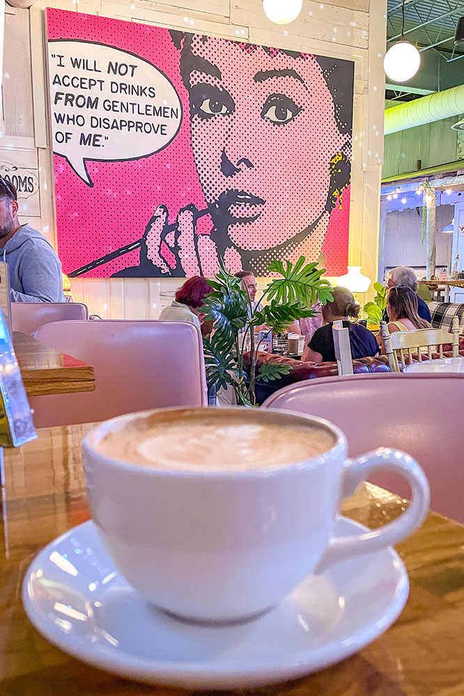 A cup of coffee drink served in a white cup on a white saucer in the foreground on a wooden table with a view of the people dining beside a white wall with a large comic artwork of woman in pink background and a thought bubble beside her. 
