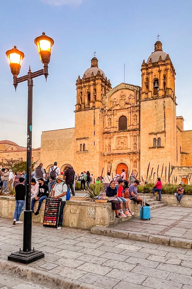 An outdoor space in front of the Santo Domingo Church with people walking, sitting and some plants beside a sidewalk with a lamp post in Oaxaca Mexico