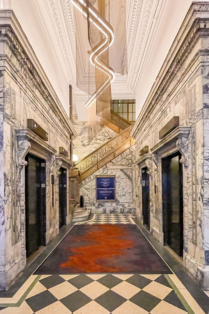 A hall with walls covered with marbled tiles and a floor of white and black tiles leading to a staircase with golden handles leading to an upper floor of the Industrialist Hotel in Pittsburgh.