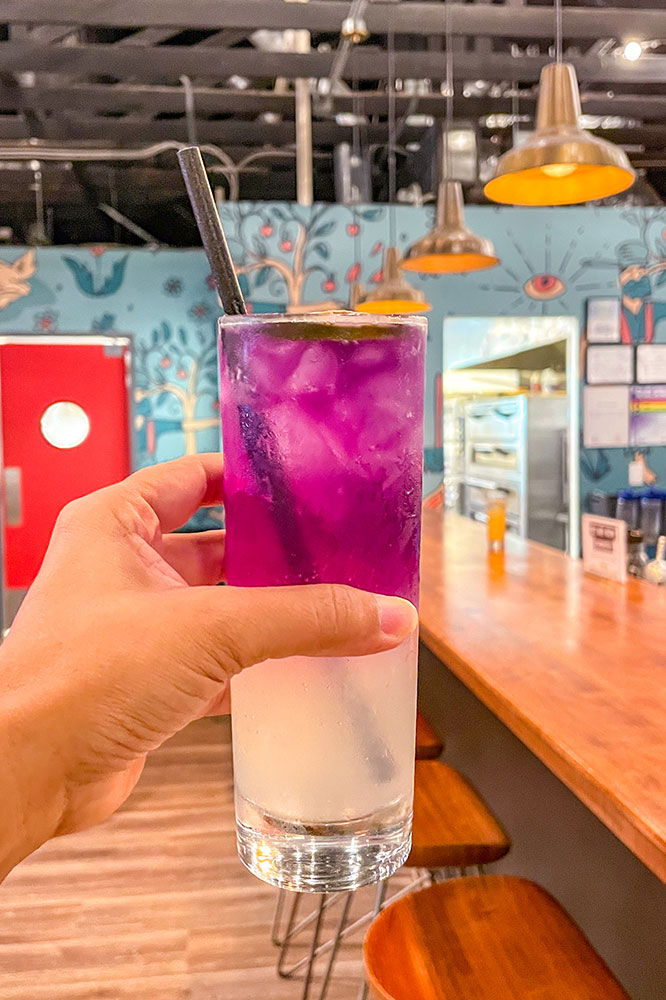 A hand holding a purple and white ice cold drink with a straw with a view of a blue base painted mural wall and a bar on the side.
