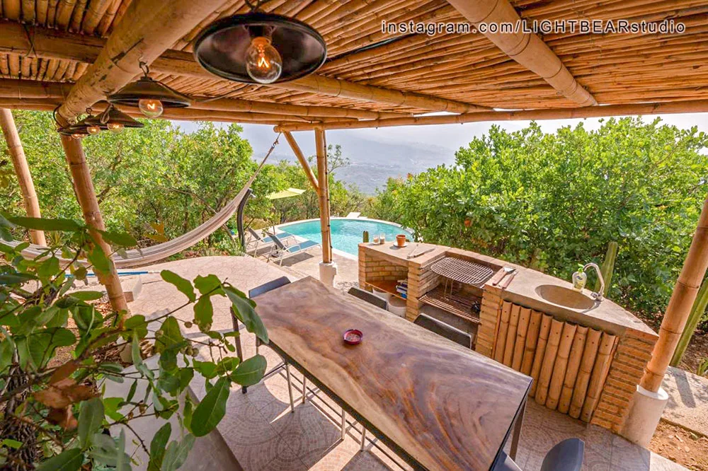 An outdoor wooden kiosk kitchen space with wooden tables and kitchen desks surrounded by tall green trees and a small blue pool in the far back in Oaxaca Mexico