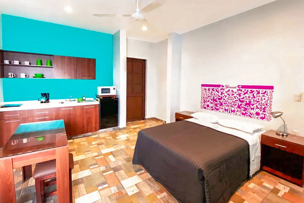 A room with a white and brown bed in between wooden side tables on brown colored tiles beside white walls, while the other half is colored in blue separating the kitchen with wooden finishings and tables and chairs.