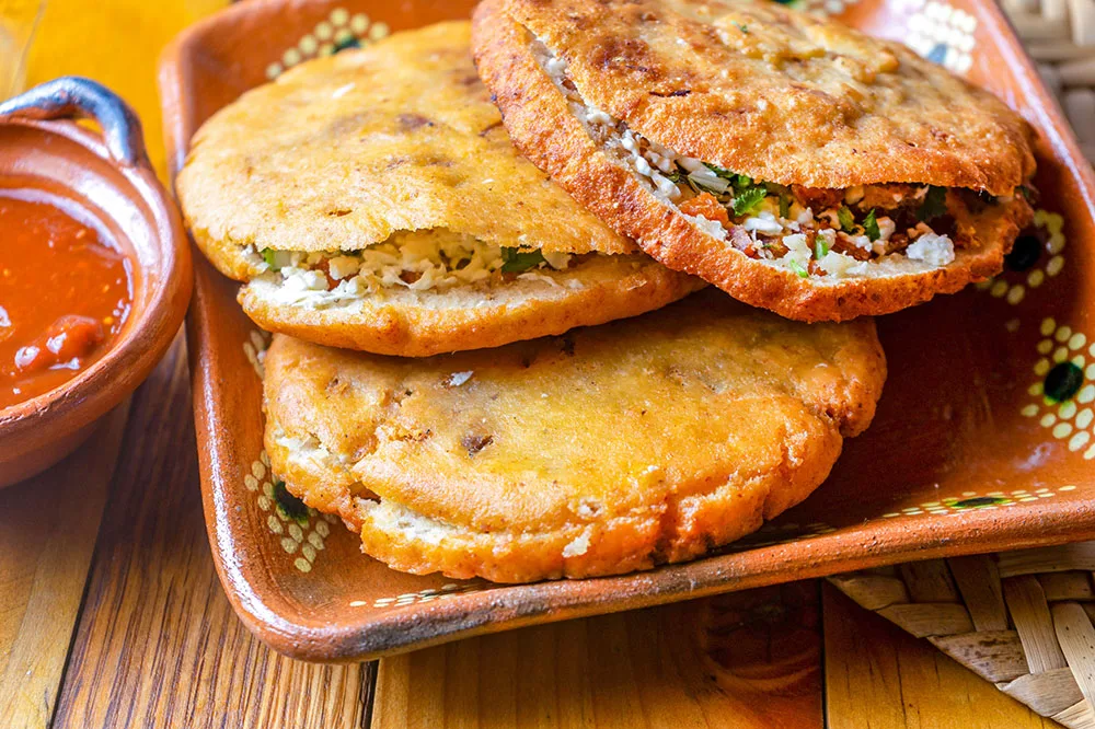 Fried tortilla pockets filled with meat, cheese, and vegetable filling served on a square deep plate with a side of a red sauce