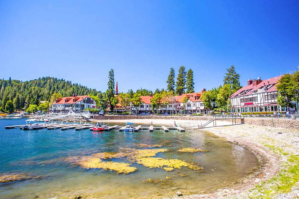 A small shore with a line of white houses while surrounded by trees in different sizes with small boats parked nearby under clear blue skies in Lake Arrowhead.
