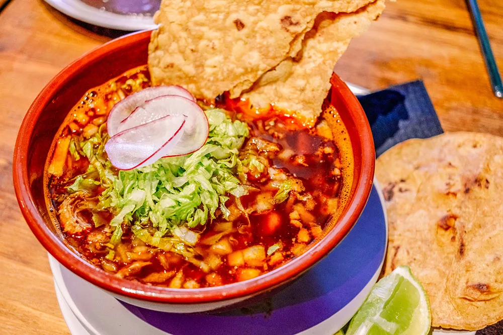 A red soup of Pozolo topped with toasted tortilla topped with cabbage and onion slices with a side of lime slices and a whole tortilla shell