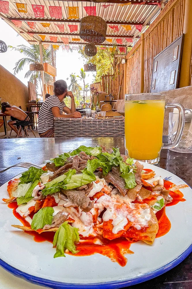 Tortilla chips covered with red sauce, melted cheese, meat slices, and vegetable slices served in a white plate served with a mug of juice