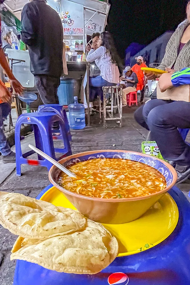 A bowl of soup with slices of vegetables and meat served with tortilla shells on a yellow plate.
