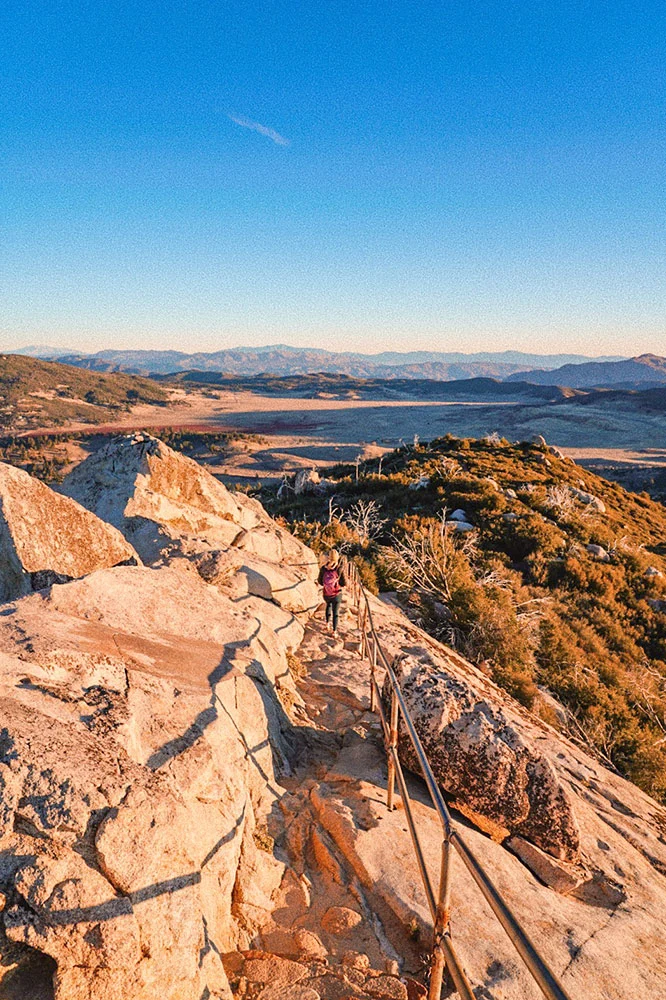 A person going down a trail while holding on a metal hand rail with a view of some bushes and fields in the background under blue skies.