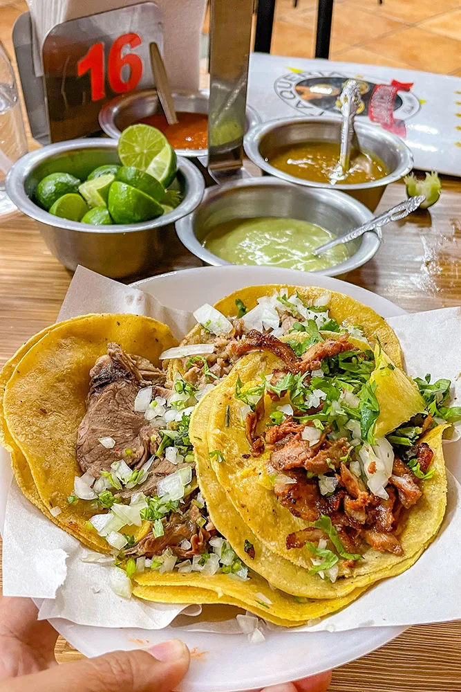 A basket of taco shells and fillings of meat, onions, and other vegetables while tin cups of sauces and lime can be found in the background.