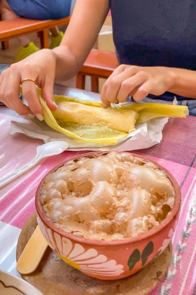 Two hands opening a Tamale, a traditional Mexican food, on table served with a cup of cocoa.