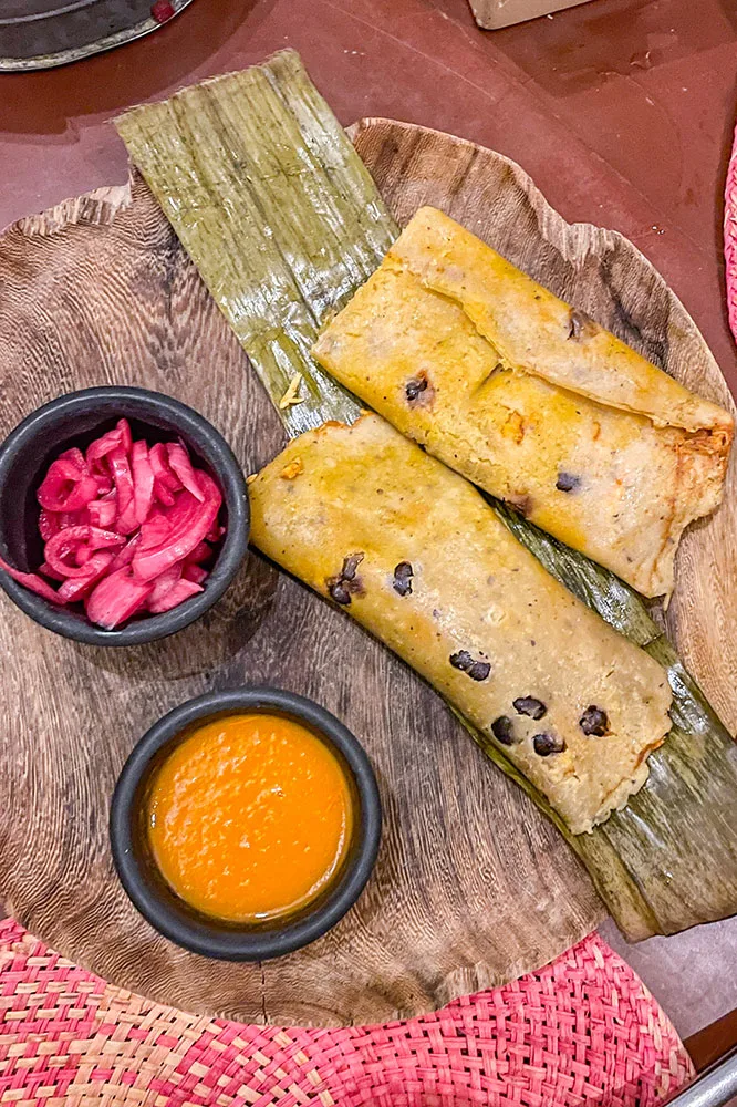 Two rolls of Tamales, one of the best traditional Mexican food, served on a wooden plate with the side of onions and an orange dip in small black cups.