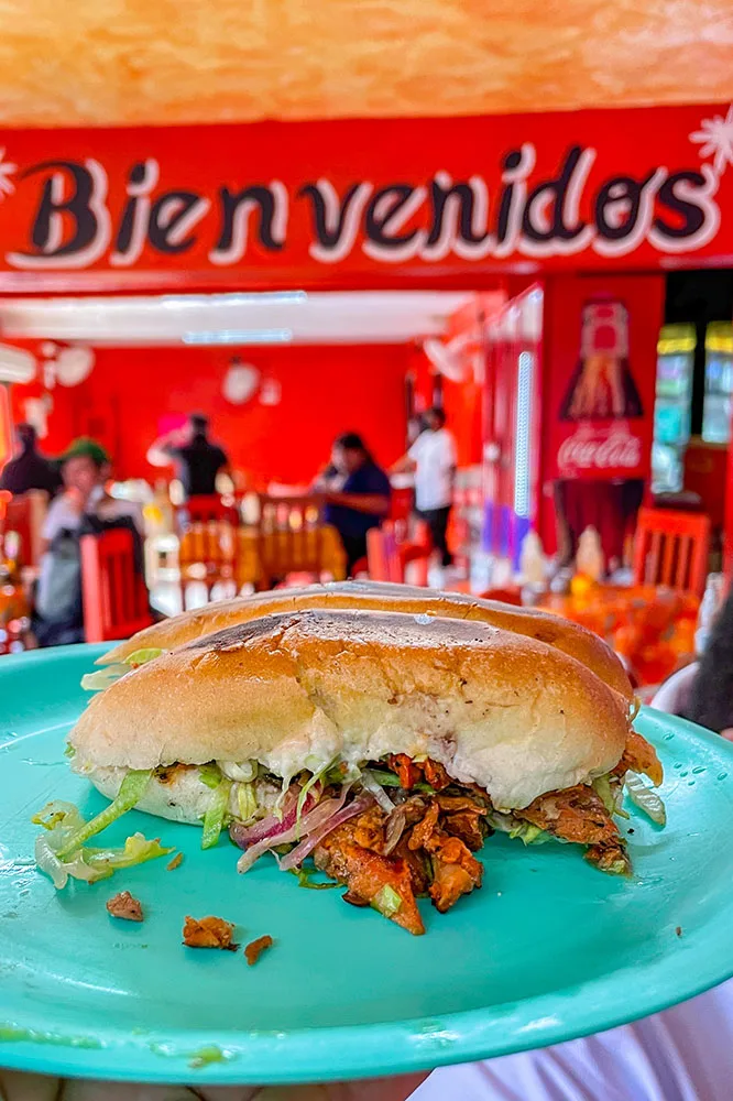 Torta, a Mexican sandwich, filled with vegetable and meat slices served on a blue plate with a background of red restaurant with red chairs and people dining and standing.