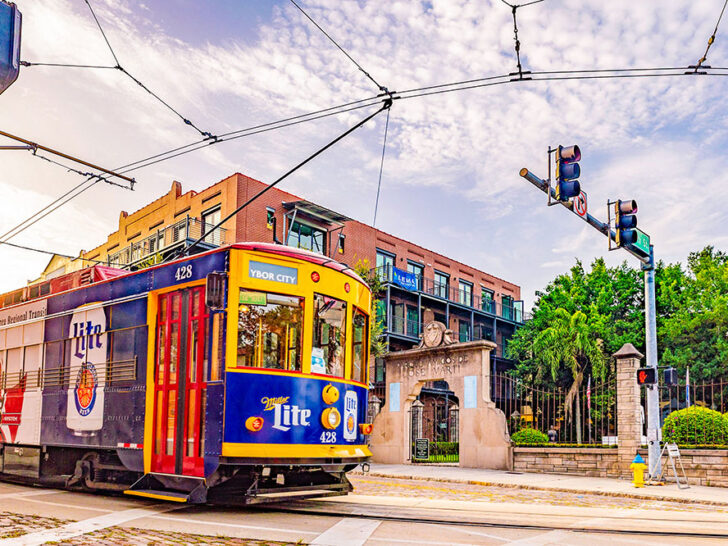 A brightly colored train of the TECO Line Streetcar System painted in blue, yellow, and red on the road with a street lamp and a building behind it under bright cloudy skies. A transportation to see hidden gems in Tampa Florida