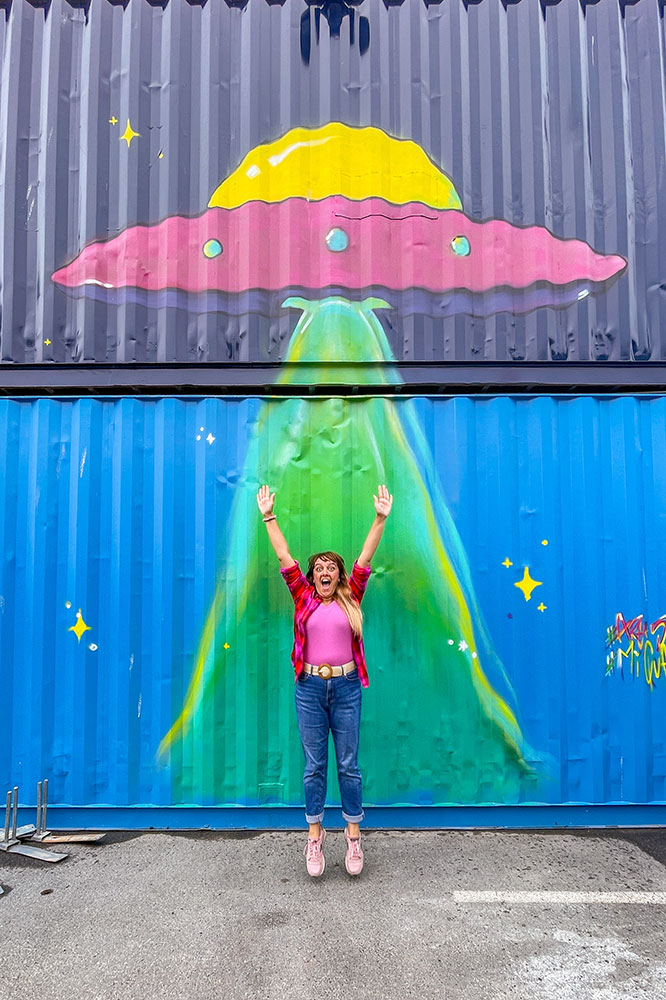 Two stacks of cargo containers with a UFO mural while I pretend to get lifted towards the spaceship.