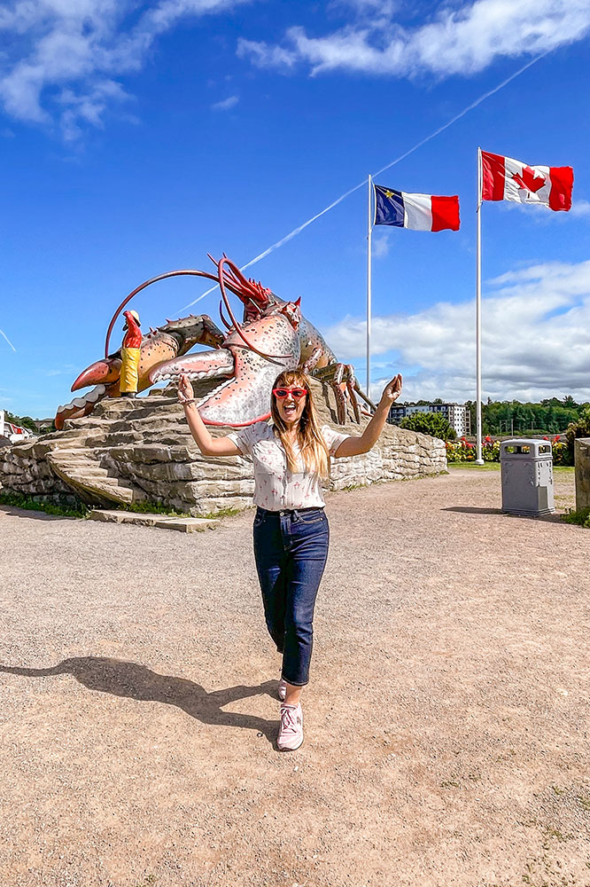 Me in a happy pose in front of the large lobster statues with the flags of Canada and Flag of Acadia waving under bright blue skies with some clouds while a garden with yellow and red flowers can be seen at the back along the Fundy Coastal Drive.