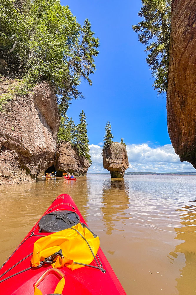 Red kayak bow with muddy waters, kayakers, rock formations, lush vegetation, clear blue skies, and clouds in the background.