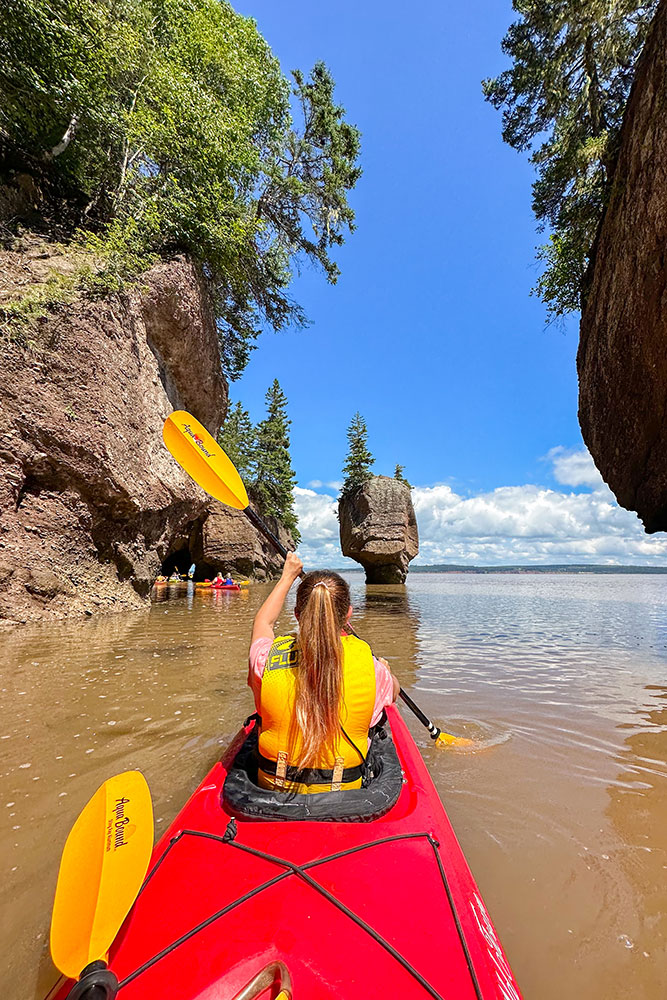 Me rowing a red kayak on the waters with the view of other people on kayaks in the back nearby the rock formations under bright blue skies with some clouds at the back along the Fundy Coastal Drive.
