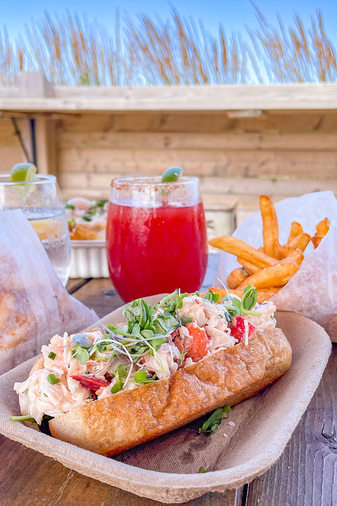 A lobster roll sandwich served with fries and some red drinks on a wooden table.