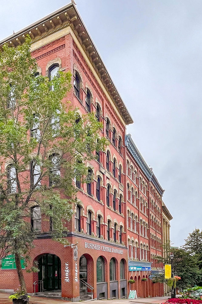 A multi-storied red-bricked building on a street in Canada under dark cloudy skies with some thin trees in the front and sides.
