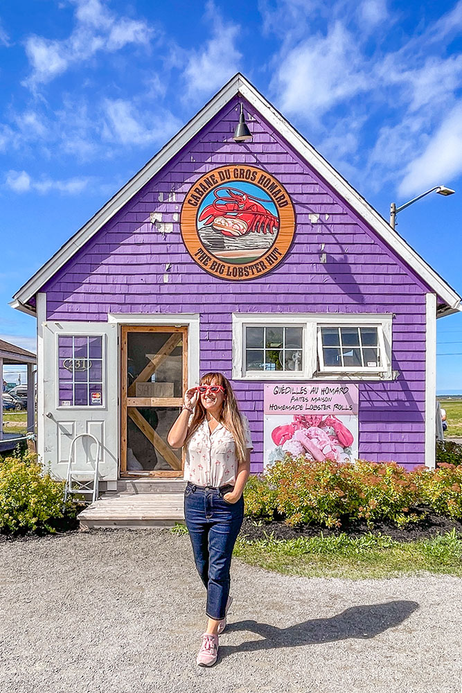 Me in front of the purple barn house building of The Big Lobster Hut with a door and two small white windows with bushes in front under bright blue skies with some clouds along the Fundy Coastal Drive.