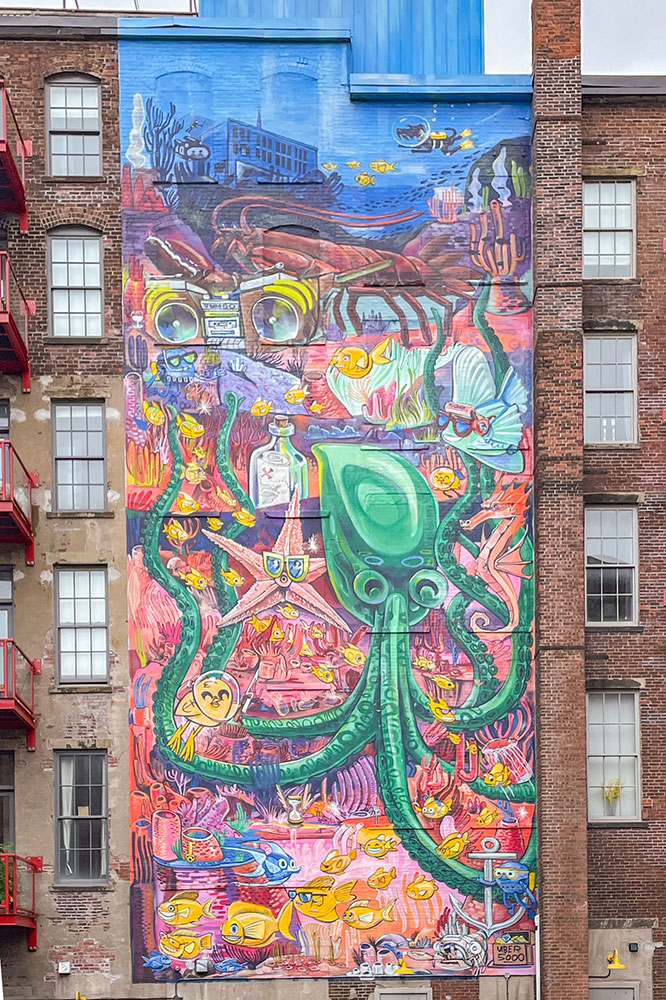A portrait view of a red-bricked building, while the middle wall features a mural of sea creatures like the octopus, types of fish, ship anchors, and bottles with a letter in them.
