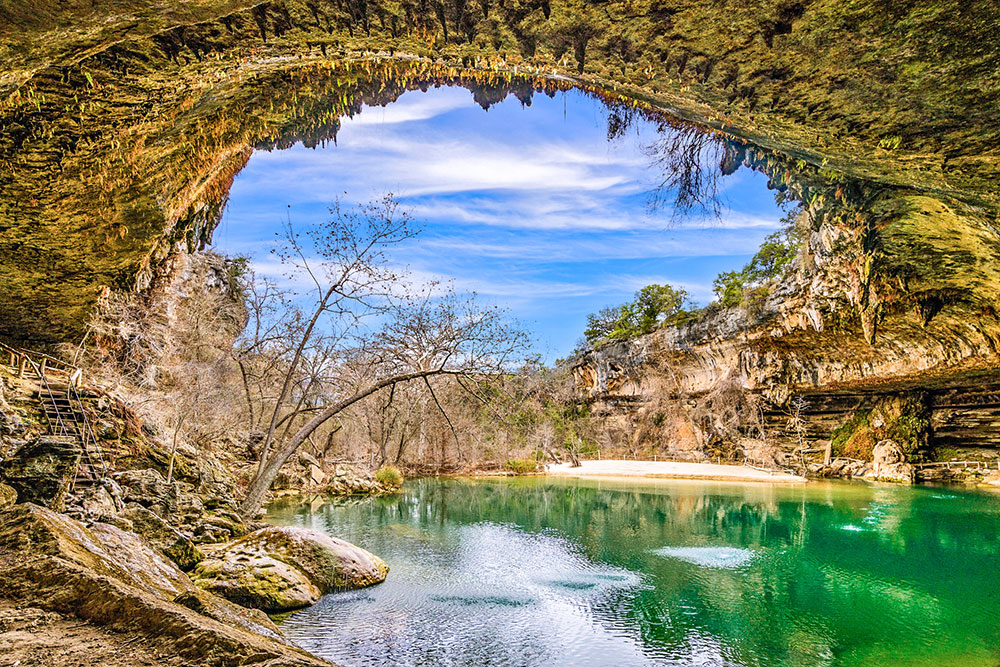 The Hamilton Pool Preserve featuring a large hole with crystal-clear green waters under bright blue skies, Texas Hill Country road trip.